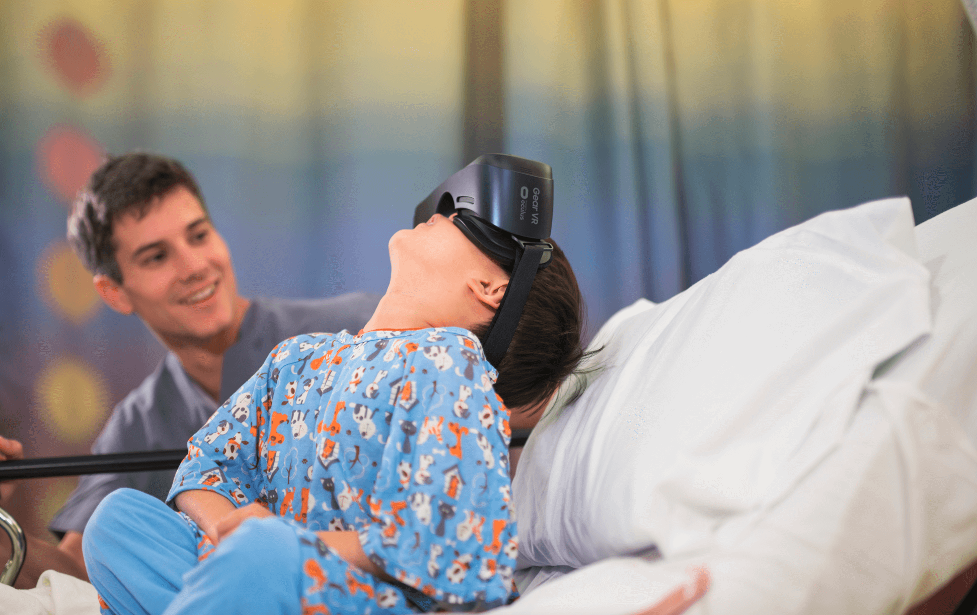 Smiling clinician beside a child in hospital pyjamas wearing a VR headset in bed