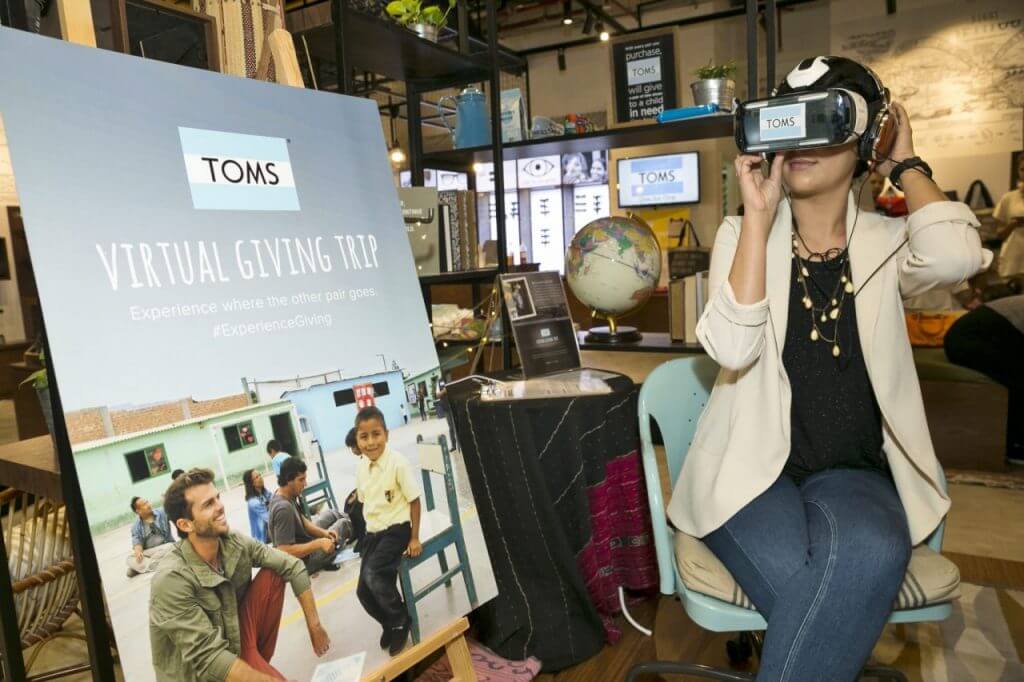 Woman in a TOMS-branded VR headset beside a Virtual Giving Trip poster in a TOMS shop